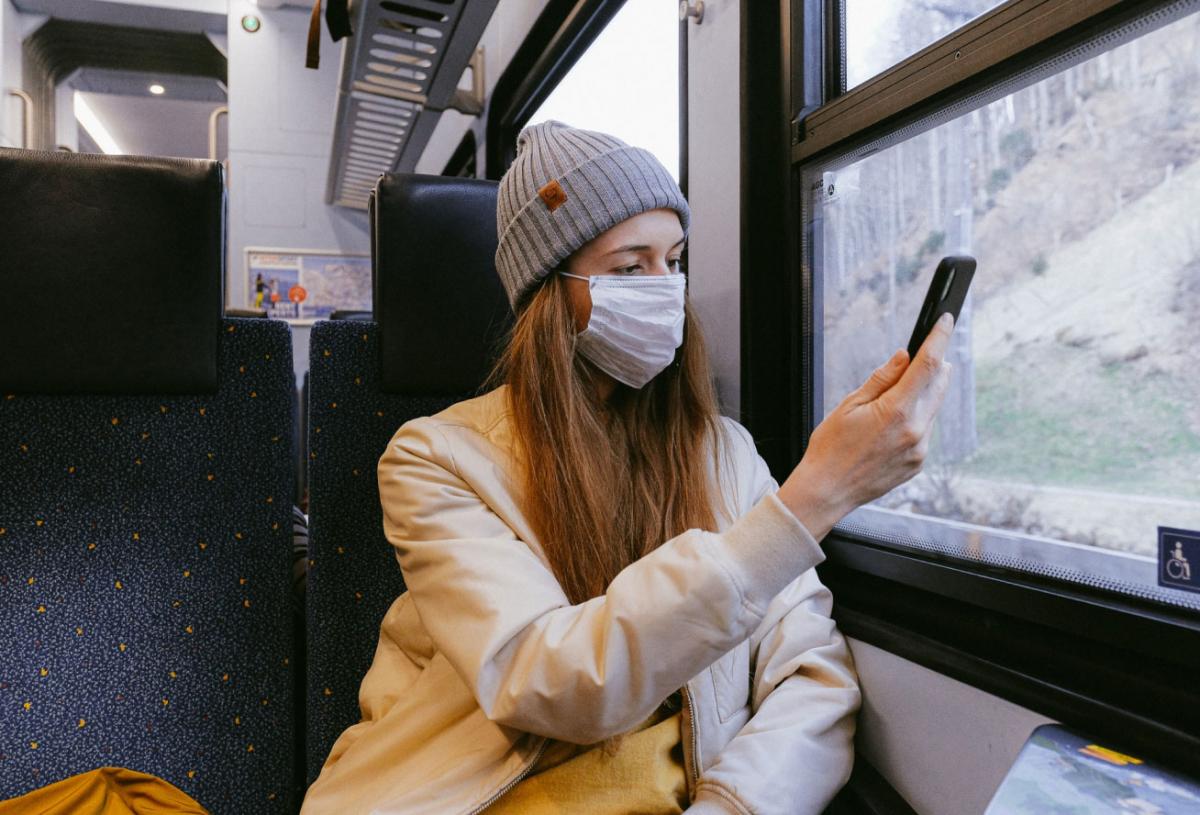 Person sits inside of a train on a rainy day wearing a coat, beanie, and mask.